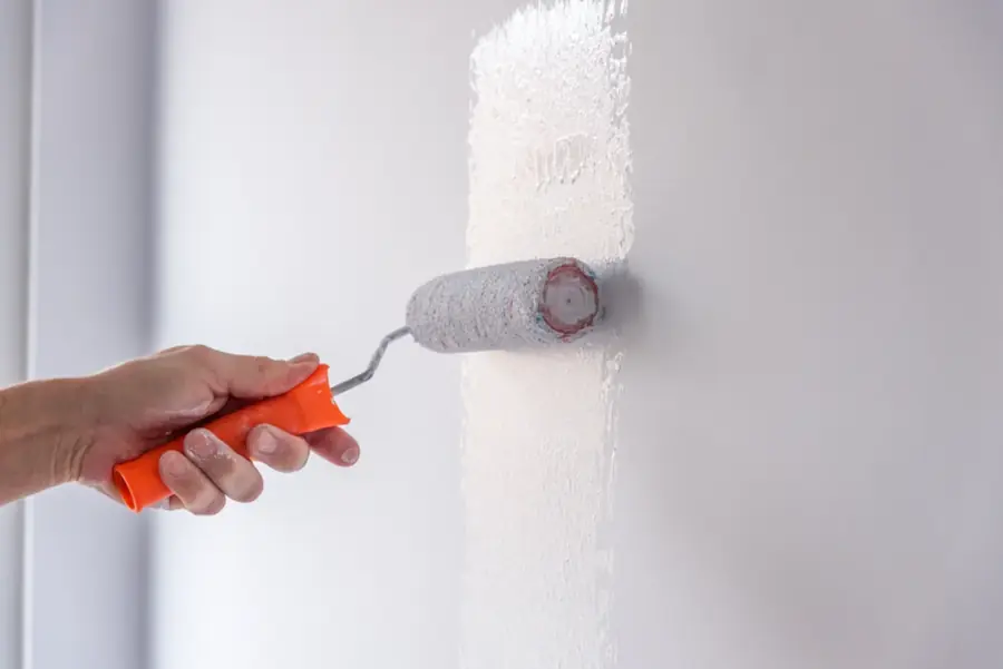 A hand holding a paint roller applying white paint to an interior wall