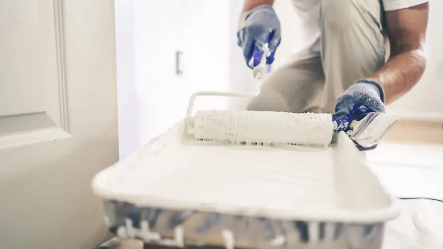 A painter rolling white paint onto an interior wall beside a window