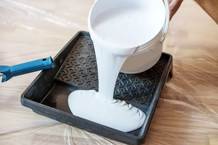 White paint being poured from a bucket into a roller tray on a drop-sheeted floor